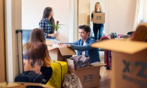 boy and girl on the floor packing their apartment with XYZ Storage moving boxes, one girl sitting on a couch holding a plant, another girl standing holding an XYZ Storage moving box